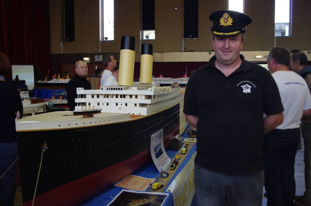 Un homme portant une casquette d'officier et un T-shirt AFT pose à côté de la grande maquette du Titanic