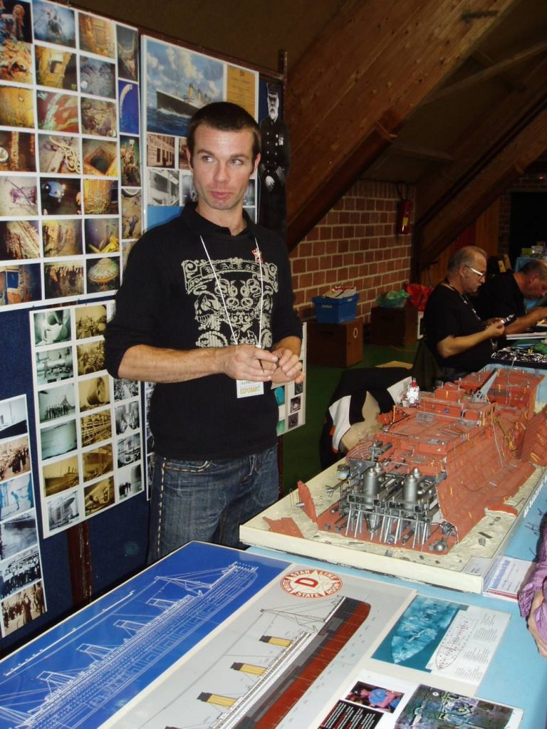 Un homme pose à côté d'une maquette de l'épave et d'images du navire.